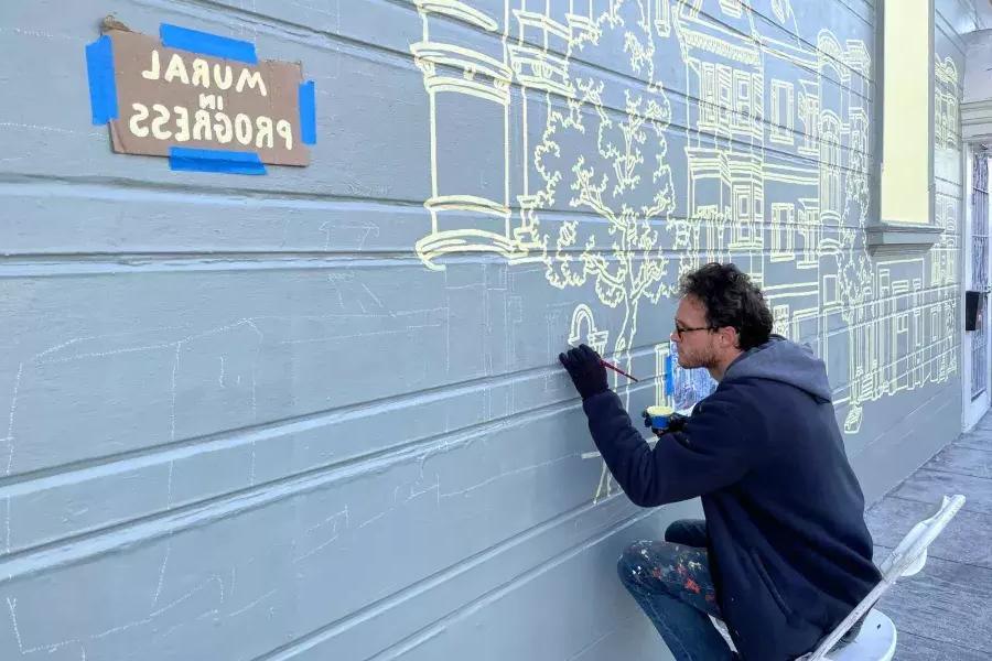 Um artista pinta um mural na lateral de um prédio no教会区, 和 uma placa colada no prédio que diz "Mural em andamento". 加州贝博体彩app.